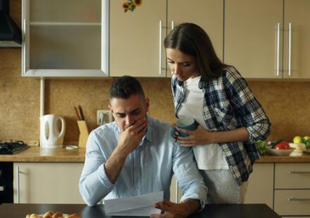 Man seated at a kitchen table reads a document with a worried expression while a woman leans over from his side, holding a mug and offering support.