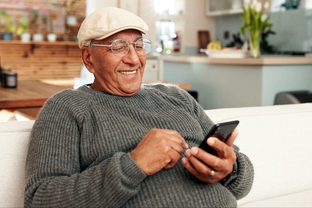 Smiling elderly man in a gray sweater and beige cap uses a smartphone on a couch at home.