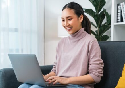 Woman in a pink sweater typing on a laptop while sitting on a sofa.