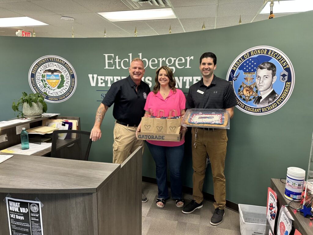Three adults stand together in a veterans center, smiling with gift bags and a cake on display in front of a teal wall with logos and a large 'Etchberger Veterans Center' sign.