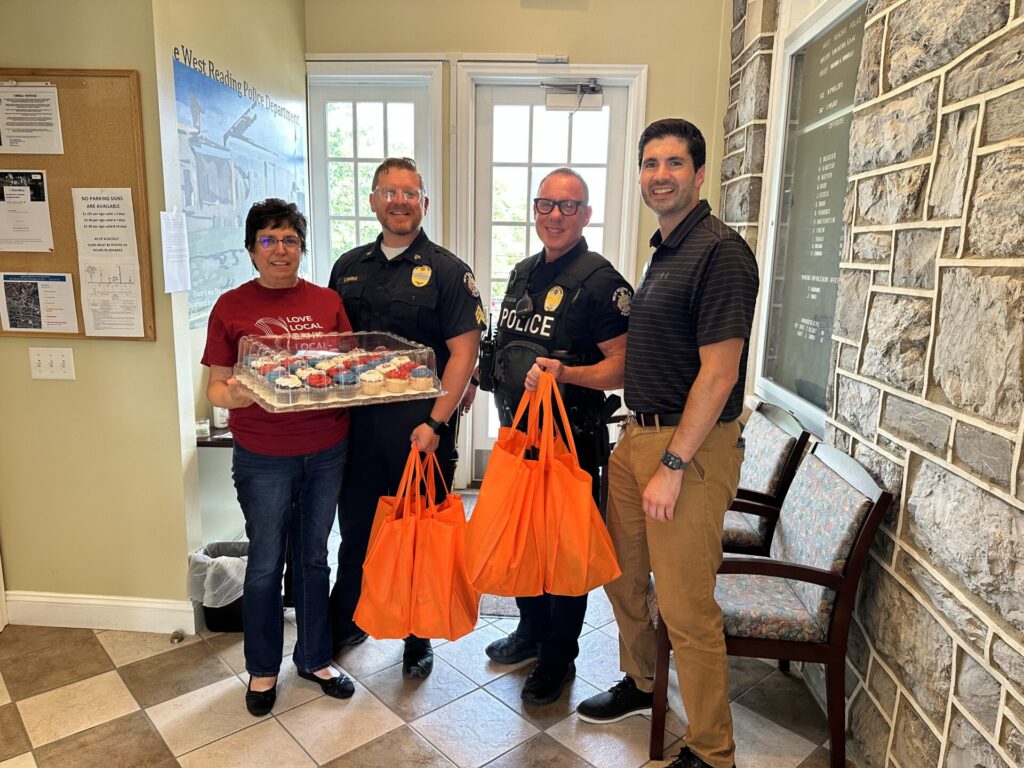 Four people stand indoors near a doorway; a woman in a red shirt holds a tray of colorful cupcakes while two uniformed police officers and a man in a striped polo smile beside her, each holding bright orange tote bags.