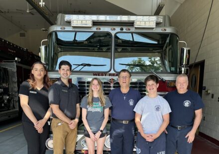 Six people—two women and four men/boys—pose in front of a large fire engine inside a fire station, smiling for a group photo.