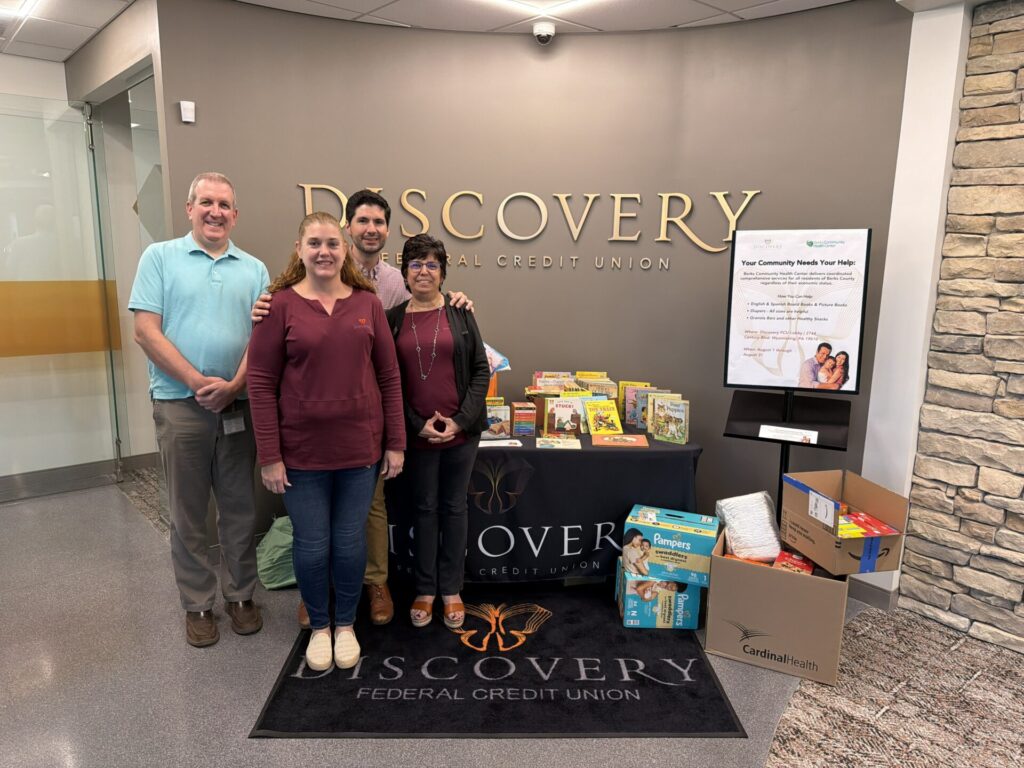 Four people stand together in a credit union lobby, posing for a group photo in front of a large 'Discovery Federal Credit Union' sign.