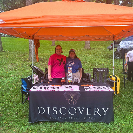 images for community involvement page | Discovery FCU Two smiling people stand behind a Discovery Federal Credit Union booth with brochures under an orange canopy in a grassy park.