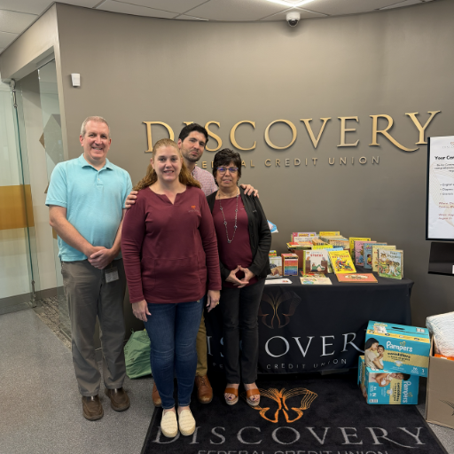 images for community involvement page 1 | Discovery FCU Four adults pose in a credit union lobby beside a display of children's books under the DISCOVERY CREDIT UNION sign.
