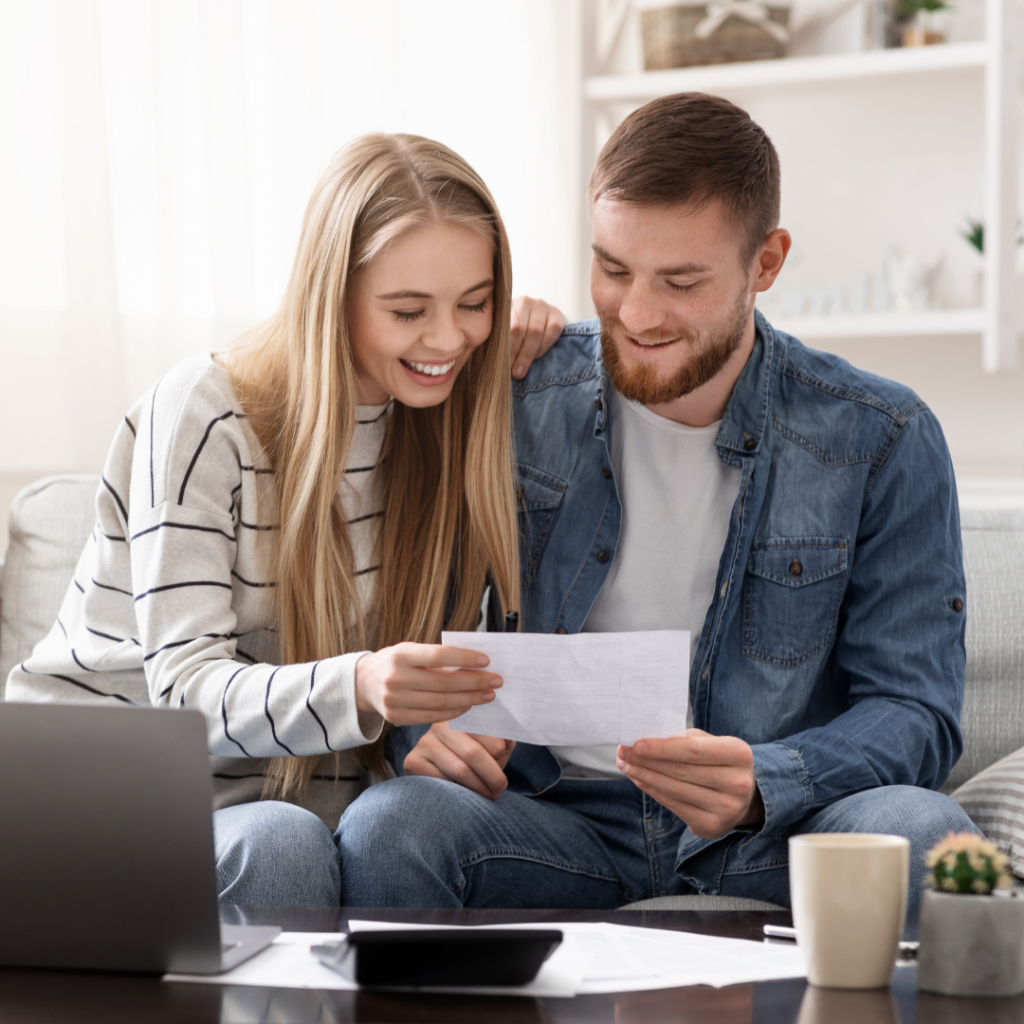 Smiling couple on a sofa reading a sheet of paper together, with a laptop on the table nearby.